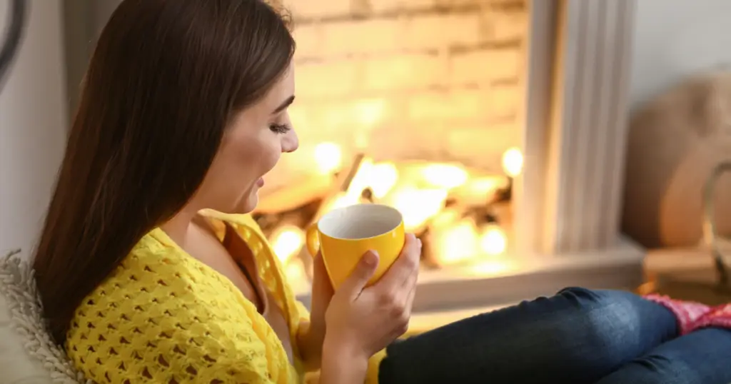 Relaxed woman enjoying a hot drink by a cozy fireplace, illustrating self-care and relaxation during wedding planning during holidays.